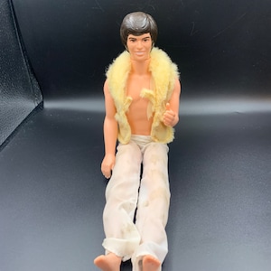 May include: A vintage doll with brown hair, wearing a white fur vest and white pants. The doll is sitting with its legs crossed and has a slight smile on its face.