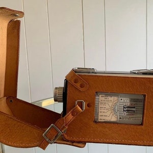 May include: A brown leather camera case with a strap and a silver camera inside. The camera has a silver dial with markings for different settings.