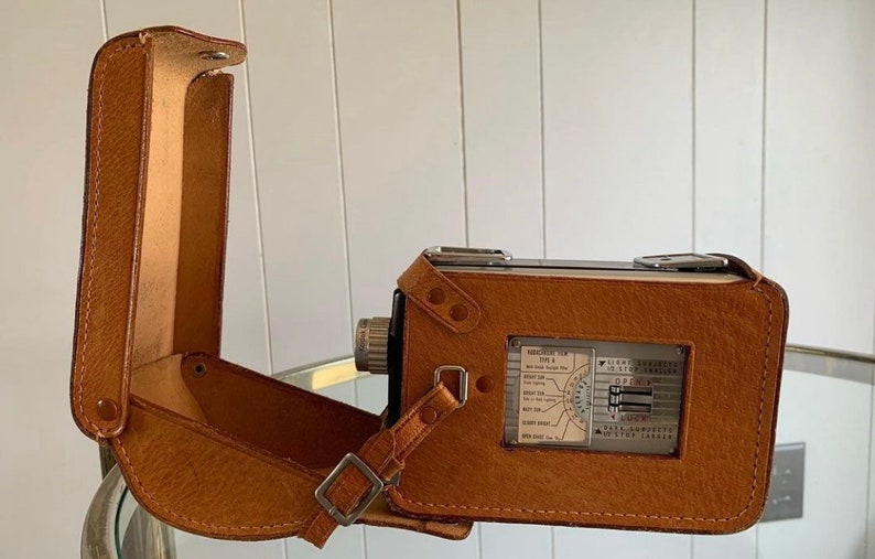 May include: A brown leather camera case with a strap and a silver camera inside. The camera has a silver dial with markings for different settings.