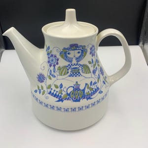 May include: A white ceramic teapot with a blue and green floral design featuring a girl sitting with a cup and teapot. The teapot has a lid and a handle.