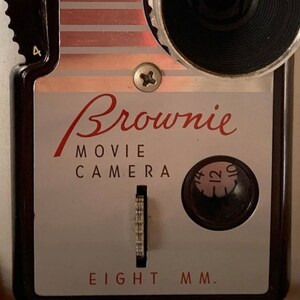 May include: A vintage silver and black Brownie Movie Camera with a red and white striped design. The camera has a black lens and a dial with numbers 10, 12, and 14. The text "Brownie Movie Camera Eight MM." is printed on the front of the camera.