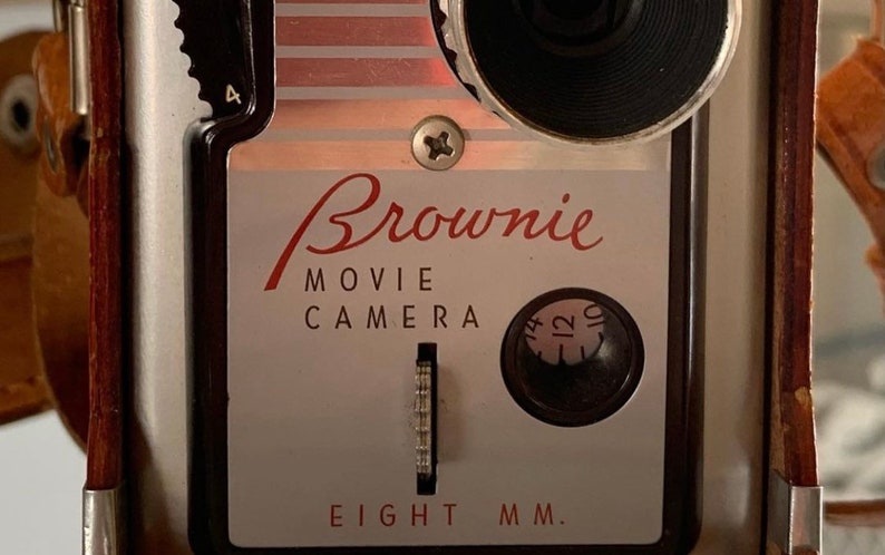 May include: A vintage silver and black Brownie Movie Camera with a red and white striped design. The camera has a black lens and a dial with numbers 10, 12, and 14. The text "Brownie Movie Camera Eight MM." is printed on the front of the camera.
