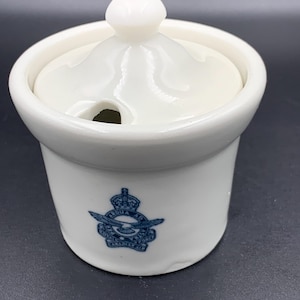 May include: A white ceramic sugar bowl with a lid and a small knob. The bowl has a blue emblem with text and a crown. The lid has a small opening for a spoon.