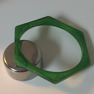 May include: A translucent green bangle bracelet with a distinct hexagonal shape. The bracelet features a marbled texture throughout the material and is positioned resting against a small, smooth, cylindrical metallic object with a reflective surface.