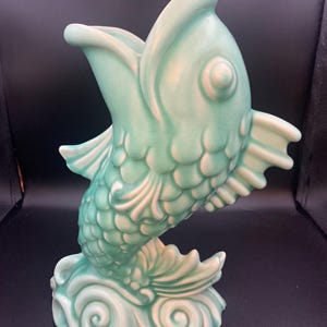 Peut inclure: Un vase en céramique en forme de poisson bondissant à la bouche ouverte. La pièce présente un vernis vert menthe clair avec des reflets blancs sur les écailles, les nageoires et la base en forme de vagues tourbillonnantes.