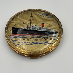 Peut inclure: Un compact doré avec un dessus rond en verre représentant une image peinte du paquebot RMS Franconia. Le navire est noir avec des accents rouges et des fenêtres blanches. L'arrière-plan est une mer bleu clair avec des nuages blancs.