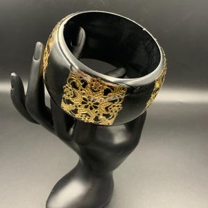 May include: A black bangle bracelet with gold filigree accents. The wide, glossy bracelet features intricate openwork designs in gold against a dark background. The jewelry is displayed on a black hand-shaped stand.