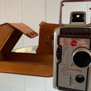 May include: A silver Kodak Brownie Movie Camera with a black leather case. The camera has a black lens and a red Kodak logo. The text "Brownie Movie Camera Eight MM" is printed on the front of the camera.