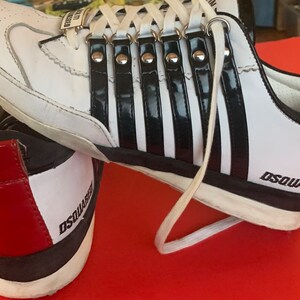 May include: White sneakers with black stripes and the word "DSQUARED" printed on the side. The shoes have white laces and a red accent on the heel. The design includes silver metal accents.