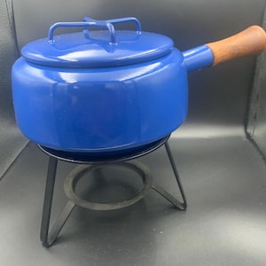 May include: A blue fondue pot with a lid and a wooden handle. The pot sits on a black metal stand with a circular base. The pot is round and has a handle on one side.