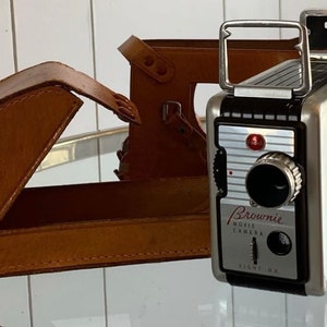 May include: A vintage silver and black Brownie Movie Camera with a leather case. The camera has a red button and the text "Brownie Movie Camera Eight MM" on the front.