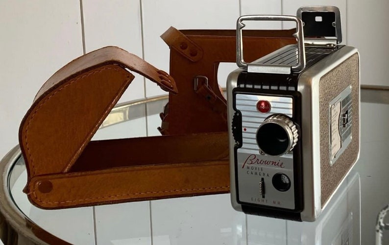 May include: A vintage silver and black Brownie Movie Camera with a leather case. The camera has a red button and the text "Brownie Movie Camera Eight MM" on the front.