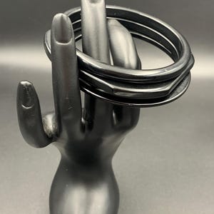 May include: A set of three smooth, solid black bangles. Two of the bangles feature a simple, rounded circular profile, while the central bangle displays a distinct hexagonal shape. The bracelets have a glossy finish and are stacked together.