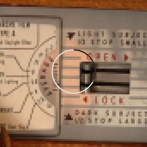 May include: A close-up of a silver control panel with a black and white dial. The dial has the words "OPEN" and "LOCK" printed on it. The panel also has the text "LIGHT SUBJECT TO STOP FALLING" and "DARK SUBJECT TO STOP LABEL".