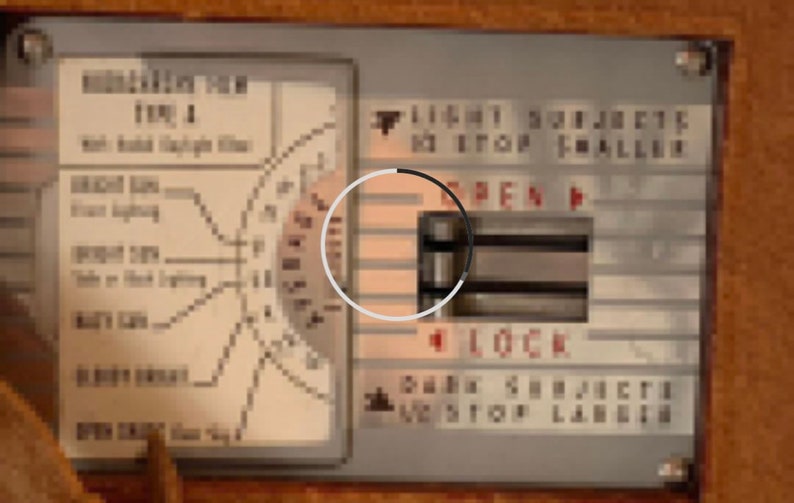 May include: A close-up of a silver control panel with a black and white dial. The dial has the words "OPEN" and "LOCK" printed on it. The panel also has the text "LIGHT SUBJECT TO STOP FALLING" and "DARK SUBJECT TO STOP LABEL".