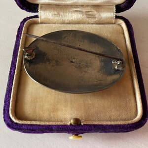 May include: A silver oval brooch with a pin on the back. The brooch is in a purple velvet box. The back of the brooch has the number "925" stamped on it.