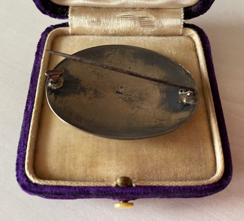 May include: A silver oval brooch with a pin on the back. The brooch is in a purple velvet box. The back of the brooch has the number "925" stamped on it.