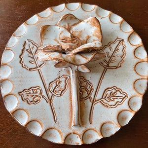 May include: A white ceramic plate with a raised, brown glazed rose and leaves. The plate has a scalloped edge with small circles around the edge.