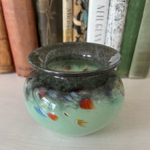 K&ouml;nnte beinhalten: Eine kleine, runde, gr&uuml;ne Glasvase mit einem dunkelgrauen Rand. Die Vase hat ein wirbelndes Muster aus Wei&szlig;, Blau und Orange.
