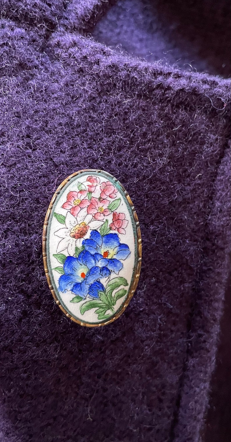 Może przedstawiać: Owalna broszka w stylu vintage z kwiatowym wzorem. Broszka przedstawia r&oacute;żowe, białe i niebieskie kwiaty z zielonymi liśćmi na białym tle. Broszka jest otoczona złotą metalową ramką.