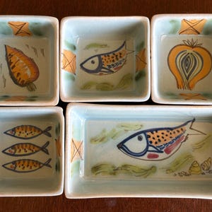 May include: A collection of six small, hand-painted ceramic dishes. Each dish has a light blue interior with a different food-themed illustration, including a carrot, fish, and onion. The dishes are square and rectangular.