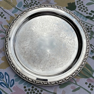 Pode incluir: Uma bandeja redonda prateada com um intrincado padrão floral gravado no centro. A bandeja tem uma borda decorativa com um design repetitivo. A bandeja é exibida em uma superfície com padrão floral.
