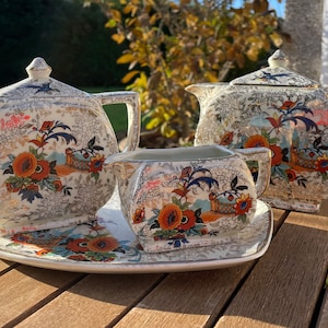 Puede incluir: Un juego de té vintage con un patrón floral en tonos naranja, azul y blanco. El juego incluye una tetera, una jarra de crema y una bandeja.