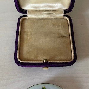 May include: A vintage brooch featuring a delicate design of two pink roses on a pale blue background. The brooch is presented in a purple velvet box.
