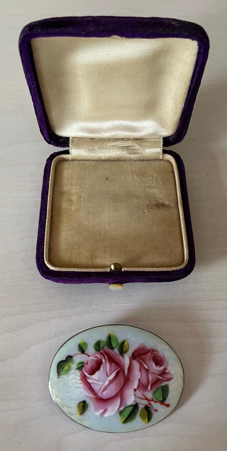 May include: A vintage brooch featuring a delicate design of two pink roses on a pale blue background. The brooch is presented in a purple velvet box.
