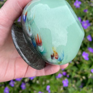 K&ouml;nnte beinhalten: Eine kleine, runde, gr&uuml;ne Glasvase mit einem gesprenkelten, dunkelgrauen Sockel. Die Vase hat eine gl&auml;nzende Oberfl&auml;che und zeigt bunte, abstrakte Muster.