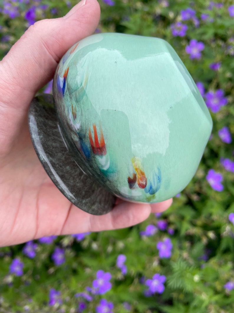 K&ouml;nnte beinhalten: Eine kleine, runde, gr&uuml;ne Glasvase mit einem gesprenkelten, dunkelgrauen Sockel. Die Vase hat eine gl&auml;nzende Oberfl&auml;che und zeigt bunte, abstrakte Muster.