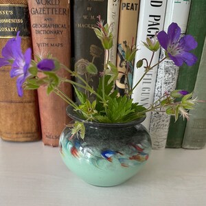 K&ouml;nnte beinhalten: Eine kleine, gr&uuml;n und schwarz glasierte Keramikvase mit floralem Muster. Die Vase ist mit einem Strau&szlig; lila Wildblumen und gr&uuml;nen Bl&auml;ttern gef&uuml;llt. Die Vase steht auf einer wei&szlig;en Oberfl&auml;che vor einer Reihe von B&uuml;chern.