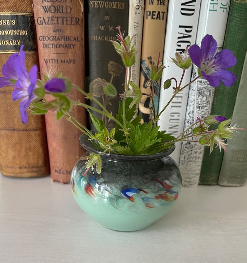 K&ouml;nnte beinhalten: Eine kleine, gr&uuml;n und schwarz glasierte Keramikvase mit floralem Muster. Die Vase ist mit einem Strau&szlig; lila Wildblumen und gr&uuml;nen Bl&auml;ttern gef&uuml;llt. Die Vase steht auf einer wei&szlig;en Oberfl&auml;che vor einer Reihe von B&uuml;chern.
