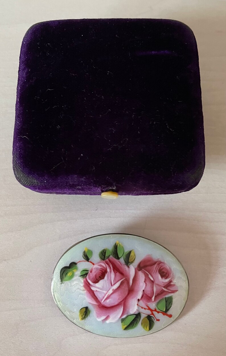 May include: A silver oval brooch with a white enamel background and hand-painted pink roses with green leaves.