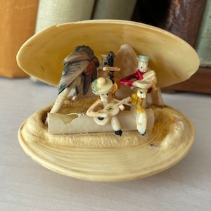 Könnte beinhalten: Eine Vintage-Musikerfigur aus drei Musikern, die Instrumente spielen, in einer Muschel. Die Musiker tragen bunte Kleidung und Hüte. Die Muschel ist weiß und hat eine strukturierte Oberfläche.