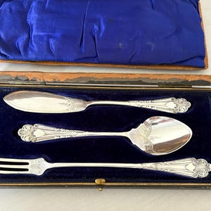 Op de afbeelding: Een zilveren botermes, lepel en vork set in een blauw fluwelen doos. Het zilverwerk heeft sierlijke bloemendetails op de handvatten en wordt gepresenteerd tegen een blauwe fluwelen achtergrond. De doos is open.