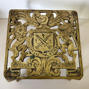 May include: An antique brass trivet with an intricate openwork design. The square trivet features a coat of arms with a lion and unicorn. The words "Dieu et mon droit" are visible. The trivet is supported by small feet. Measures approximately 15 cm x 15 cm.