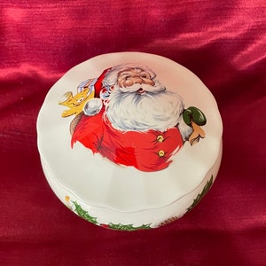 Op de afbeelding: Witte keramische kerstversieringendoos met een illustratie van een rode en witte kerstman die een cadeau en een teddybeer vasthoudt. De doos is versierd met groene hulstbladeren.