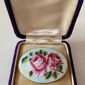 May include: A silver oval brooch with a hand-painted design of two pink roses with green leaves on a white background. The brooch is in a purple velvet box.
