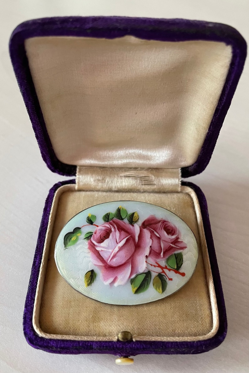 May include: A silver oval brooch with a hand-painted design of two pink roses with green leaves on a white background. The brooch is in a purple velvet box.