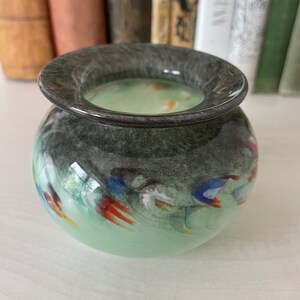 K&ouml;nnte beinhalten: Eine kleine, runde, gr&uuml;ne Glasvase mit einem dunkelgrauen Rand. Die Vase hat ein wirbelndes Muster aus Wei&szlig;, Blau und Rot.