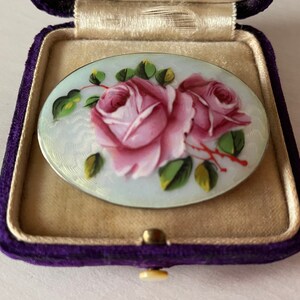 May include: A vintage oval-shaped brooch featuring a delicate hand-painted design of two pink roses with green leaves on a white background. The brooch is set in a silver tone metal.