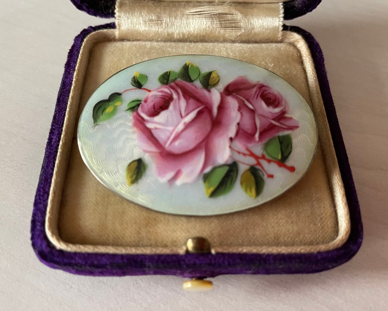 May include: A vintage oval-shaped brooch featuring a delicate hand-painted design of two pink roses with green leaves on a white background. The brooch is set in a silver tone metal.