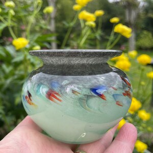 K&ouml;nnte beinhalten: Eine kleine, runde, hellgr&uuml;ne Glasvase mit einem dunkelgrauen Rand. Die Vase hat ein wirbelndes Muster aus bunten Glassplittern, darunter Rot, Orange, Blau und Gr&uuml;n.