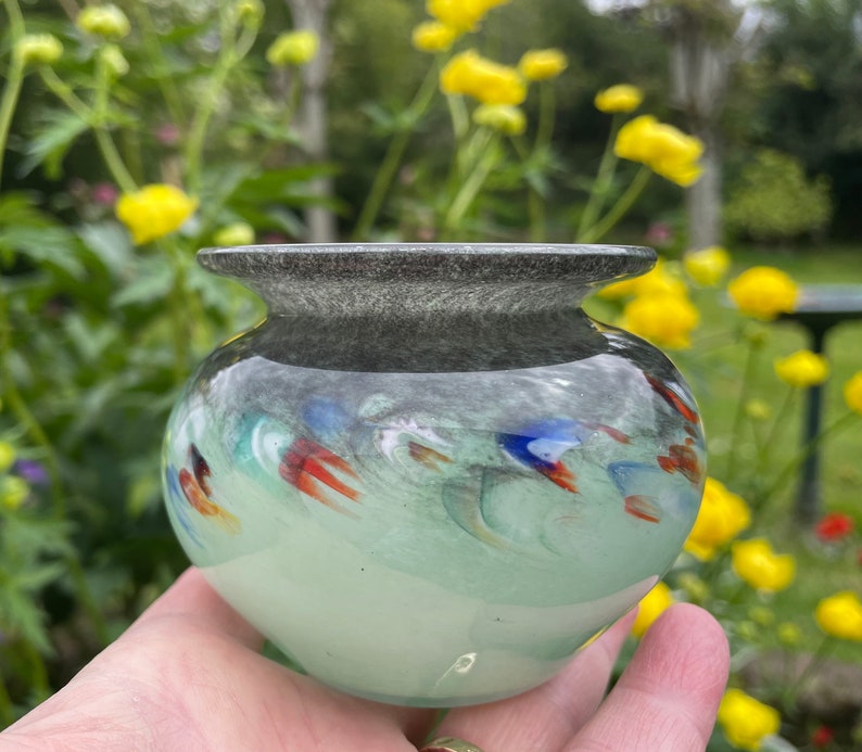 K&ouml;nnte beinhalten: Eine kleine, runde, hellgr&uuml;ne Glasvase mit einem dunkelgrauen Rand. Die Vase hat ein wirbelndes Muster aus bunten Glassplittern, darunter Rot, Orange, Blau und Gr&uuml;n.