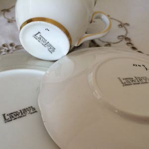 Może przedstawiać: Biały zestaw filiżanki i spodeczka do herbaty z pozłacanym wykończeniem. Filiżanka i spodeczek są oznaczone tekstem "China Glass Lawleys Stoke-on-Trent, Staffs."