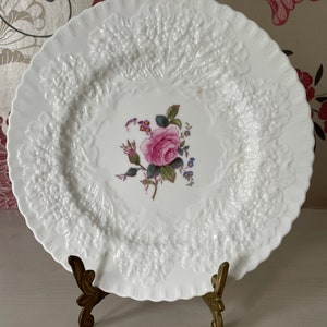 May include: White porcelain plate with a raised, embossed design. The center features a pink rose with green leaves and pink and blue flowers.