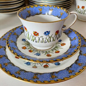 Op de afbeelding: Een blauwe en witte theekop en schotelset met een bloemenpatroon. De kop en schotel hebben een gouden rand en bevatten een ontwerp met kleurrijke bloemen en groene bladeren.