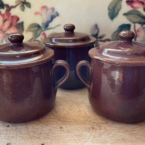 May include: Three brown ceramic lidded pots with handles. The pots are all the same size and shape, and they have a glossy finish.