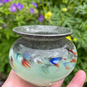 K&ouml;nnte beinhalten: Eine kleine, runde, gr&uuml;ne Glasvase mit einem dunkelgrauen Rand. Die Vase hat ein wirbelndes Muster aus Wei&szlig;, Blau und Orange.